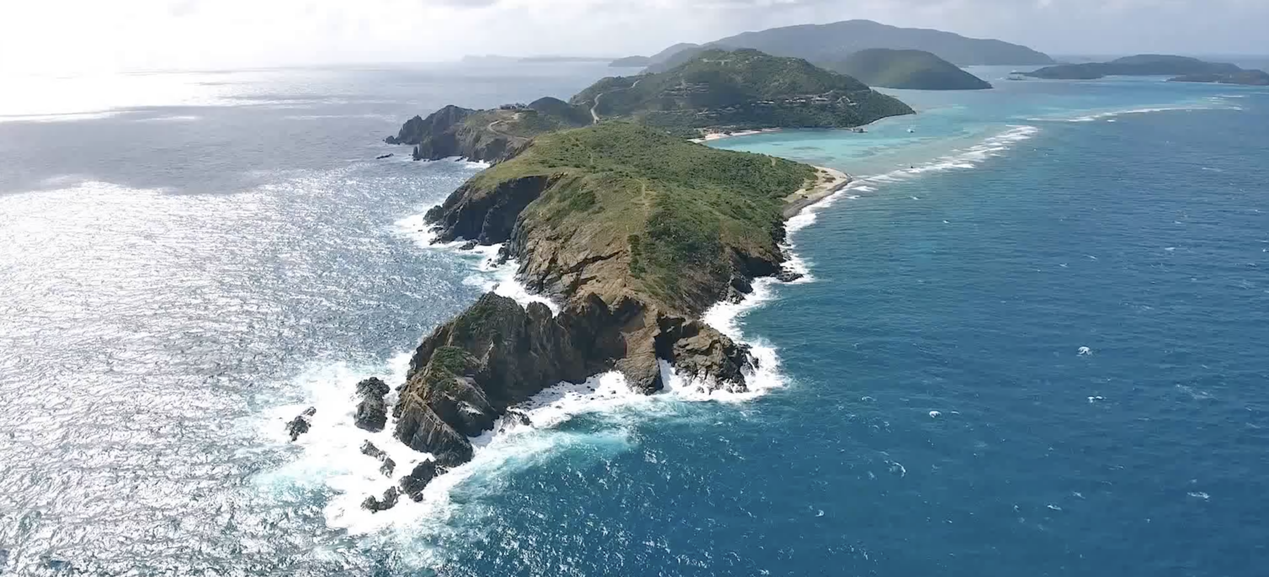 Aerial view of the rugged, narrow Pajaros Point peninsula with steep cliffs and lush green vegetation, surrounded by clear blue ocean waters. Waves crash against the coastline, and distant islands can be seen on the horizon under a partly cloudy sky, marking this scenic neighborhood.